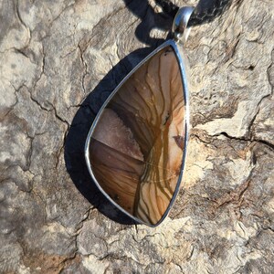 May include: A silver pendant with a brown and white agate stone. The stone has a unique pattern of swirls and lines. The pendant is hanging from a black cord.