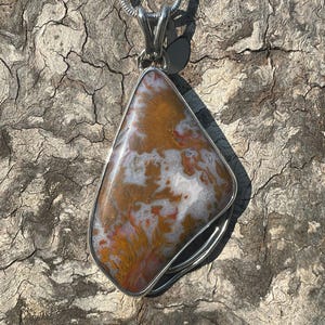 May include: A pendant necklace featuring a large, triangular stone with a silver-colored metal frame. The stone has a marbled pattern of brown, orange, white, and red hues. The necklace chain is a thin, silver-colored snake chain.