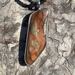 May include: A pendant necklace featuring a large, irregularly shaped stone with a silver-colored metal frame. The stone displays a mix of brown, tan, and cream colors. The necklace has a black braided cord.