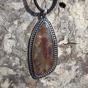 May include: A pendant necklace featuring a large, irregularly shaped stone with a silver-colored bezel. The stone has a mix of brown, red, and tan colors. The necklace has a black cord.