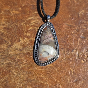 May include: A pendant featuring an irregular, polished stone with swirling patterns of brown, gray, and white, encased in a silver setting with a beaded border. The pendant hangs from a black cord necklace, showcasing a metallic finish.
