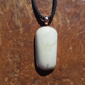 May include: A smooth, off-white, rectangular stone pendant with rounded corners, encased in a copper-toned metal setting. The pendant hangs from a black braided cord necklace, featuring a simple copper-toned bail at the top of the stone.