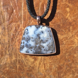 May include: A trapezoidal pendant featuring a white and gray mottled stone with dark inclusions, encased in a copper-toned metal frame. The pendant hangs from a black braided cord, creating a contrast between the metallic setting and the stone.