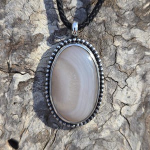 May include: Oval pendant necklace featuring a polished, light gray stone with white veining, set in a silver-toned frame with a beaded edge. The pendant hangs from a black, braided cord. The background is a textured, weathered wood surface.