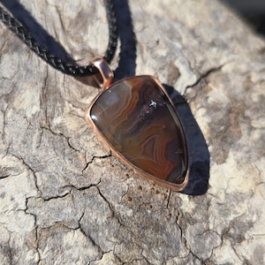 May include: A pendant featuring a polished, teardrop-shaped brown and tan banded agate stone set in a smooth copper bezel. The pendant is attached to a black braided cord necklace, showcasing intricate natural swirling patterns within the gemstone.