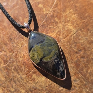 May include: A pendant featuring a polished, diamond-shaped stone with swirling patterns of olive green, black, and grey, encased in a copper-toned metal frame. The pendant hangs from a black braided cord, creating a contrast between the dark stone.
