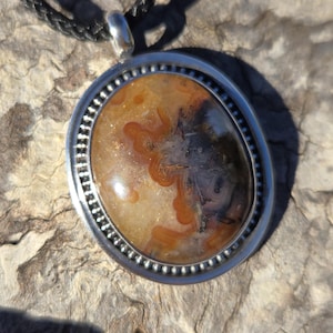 May include: A silver pendant with a brown and white agate stone set in a silver bezel. The pendant is on a black cord.