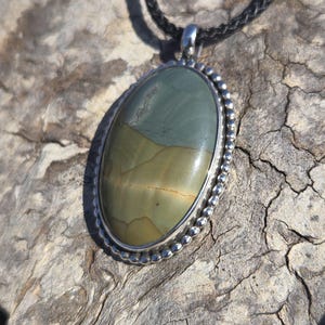 May include: A silver oval pendant with a green, brown, and white stone set in a beaded border. The pendant is hanging from a black cord.