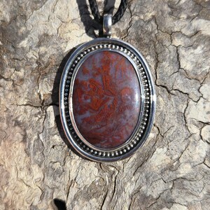 May include: A silver oval pendant with a red and white agate stone. The pendant is on a black cord.