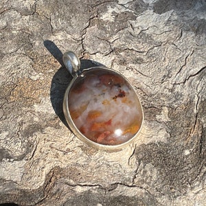 May include: A pendant with a polished, oval-shaped stone in a silver setting. The stone features a mix of colors, including white, brown, and orange. The pendant has a small loop at the top for a chain. The pendant is displayed on a textured, wooden surface.