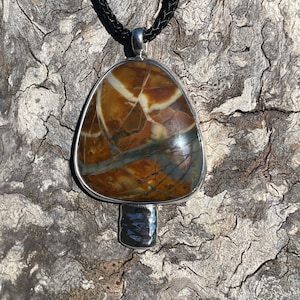 May include: A silver pendant necklace with a brown, white, and gray agate stone. The stone is set in a silver bezel and the pendant hangs from a black braided cord.