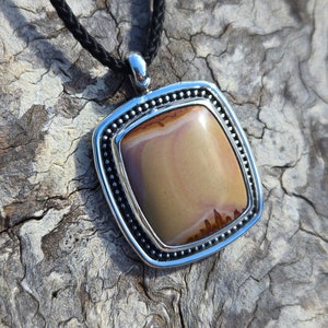 May include: A silver pendant with a brown and white agate stone set in a beaded frame. The pendant is hanging from a black braided cord.