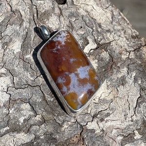 May include: A brown and white agate pendant with a silver setting. The pendant is rectangular in shape and has a smooth, polished surface.