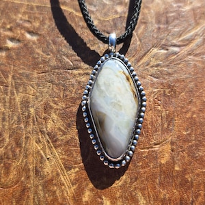 May include: A pendant necklace featuring an elongated, diamond-shaped stone with swirling cream, white, and brown patterns. The stone is set in a silver-toned metal frame with a beaded edge and hangs from a thin, black braided cord necklace.