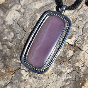 May include: A silver pendant with a rectangular purple agate stone set in a beaded border. The pendant is hanging from a black cord.