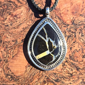 May include: A black and gold teardrop-shaped pendant with a silver beaded border hangs from a black cord. The pendant is made of a natural stone with a unique pattern.