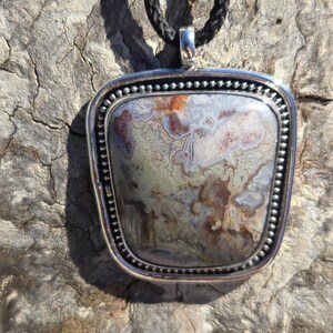 May include: A silver pendant with a square-shaped agate stone. The stone has a brown, white, and gray pattern. The pendant is on a black cord.