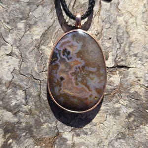 May include: A pendant necklace featuring an oval-shaped, multi-colored agate stone with intricate brown, gray, and translucent white patterns. The stone is set in a smooth copper-toned metal bezel and hangs from a black braided cord necklace.