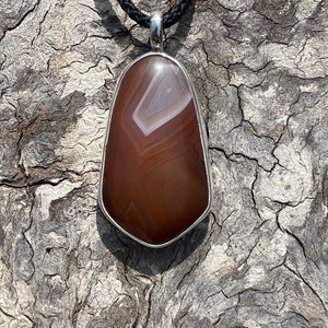 May include: A brown and white agate pendant with a silver setting, hanging from a black cord.