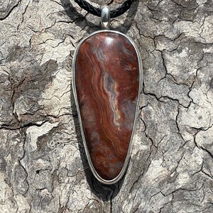 May include: A teardrop-shaped pendant with a red, brown, and white agate stone set in a silver bezel. The pendant is hanging from a black cord.