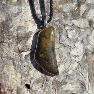 May include: A green, brown, and orange stone pendant with a silver bezel setting. The pendant is hanging from a black cord.