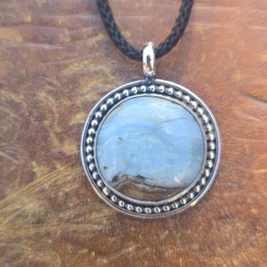 May include: A round pendant featuring a smooth, light blue and brown stone set in a silver frame with a beaded edge. The pendant hangs from a black braided cord. The stone displays swirling patterns of pale blue, white, and dark brown earth tones.