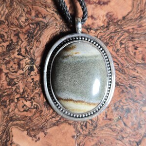 May include: A silver oval pendant with a grey and brown agate stone set in the center. The pendant is on a black cord.