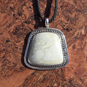 May include: A silver pendant with a white and gray stone set in a beaded border. The pendant is hanging from a black cord.