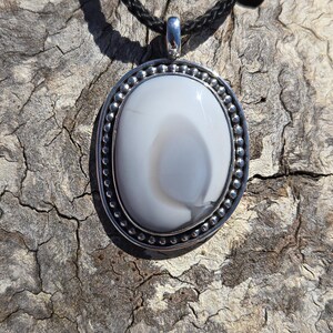 May include: A white agate pendant with a silver bezel setting, hanging from a black cord necklace.