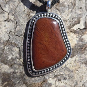 May include: A silver pendant with a red-brown stone set in a beaded border. The pendant is on a black cord.