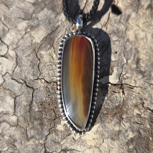 May include: A pendant featuring a polished, elongated agate stone with horizontal bands of brown, orange, and cream colors. The stone is set in a silver frame with a beaded edge and hangs from a black braided cord necklace.