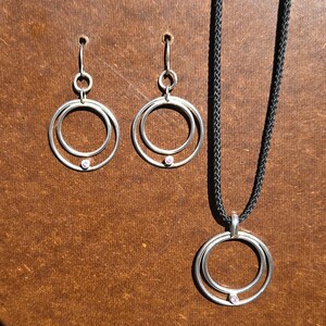 May include: A set of silver jewelry featuring a necklace and earrings. The necklace and earrings have a double circle design with a small pink gemstone in the center.