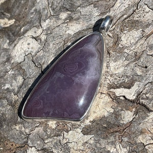 May include: A silver pendant featuring a purple and white agate stone with a swirling pattern. The pendant is shaped like a triangle and has a bail for attaching a chain.