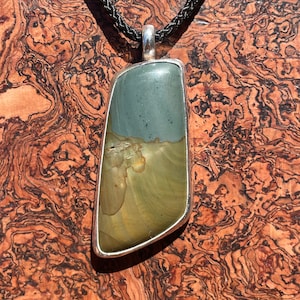May include: A silver pendant with a green and brown stone set in a silver frame. The pendant is hanging from a black cord.