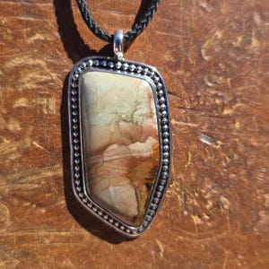 May include: A pendant necklace featuring an irregular, multi-colored stone with shades of tan, cream, and reddish-brown. The stone is set in a silver-toned metal frame with a beaded edge, suspended from a black braided cord necklace.