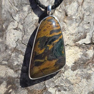 May include: A silver pendant with a brown, yellow, and green tiger eye stone. The pendant is on a black cord.