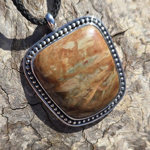 May include: A brown and beige stone pendant with a silver beaded border, hanging from a black cord.