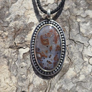 May include: A silver oval pendant with a brown and white agate stone set in a beaded border. The pendant is hanging from a black cord.