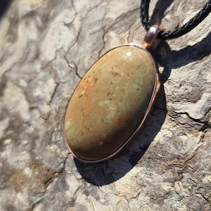May include: An oval-shaped pendant featuring a smooth, polished stone with earthy tones of tan, olive green, and brown, accented by small reddish-brown speckles. The stone is set in a copper-colored metal frame and hangs from a black braided cord.