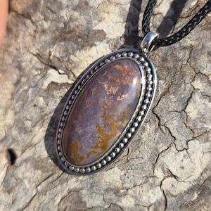 May include: A silver pendant with a brown and purple agate stone set in a beaded oval frame. The pendant is hanging from a black cord.