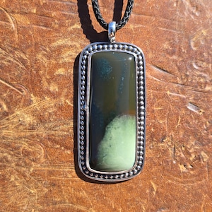 May include: A rectangular pendant featuring a smooth, dark green and black stone set in a silver frame with a beaded border. The pendant hangs from a black braided cord, showcasing a polished finish and a distinct color gradient within the stone.