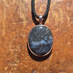 May include: An oval pendant featuring a dark, mottled stone with black and grey dendritic patterns set in a smooth copper-toned metal frame. The pendant hangs from a black braided cord, creating a contrast between the dark stone and the metal.