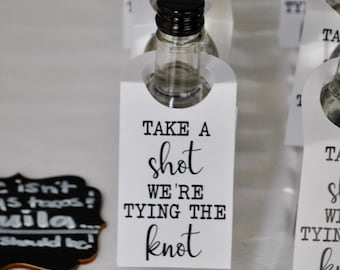 May include: White favor tags with black text that reads "Take a shot we're tying the knot". The tags are attached to small clear glass bottles.