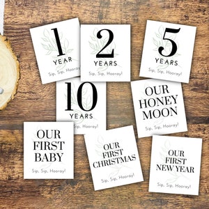 May include: Set of nine white cards with black text and green foliage illustrations. The cards feature different milestones, including "1 Year", "2 Years", "5 Years", "10 Years", "Our First Baby", "Our First Christmas", "Our First New Year", and "Our Honeymoon". Each card includes the phrase "Sip, Sip, Hooray!"