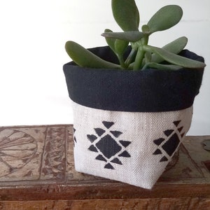 Peut inclure: Un petit pot de plante avec un bord noir et un corps de couleur crème. Le corps de couleur crème présente un motif géométrique noir répétitif. Le pot contient une plante succulente verte. Le pot est sur une surface en bois.