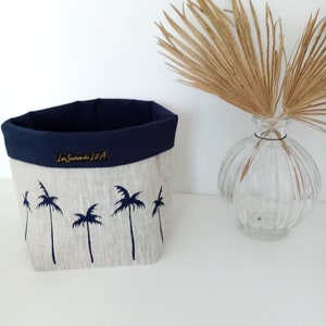 Peut inclure: Un panier de rangement en lin avec un intérieur et une bordure bleu marine. L'extérieur présente un tissu beige clair avec un motif de palmiers bleu foncé. Le texte "Les Sourires de Lil'A" est brodé en or sur le devant.