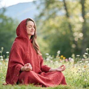 May include: A long-sleeved, hooded garment in a deep rust-red color, featuring a loose, flowing silhouette and a single visible wooden button on the side cuff. The fabric has a visible textured weave, and the hood is draped over the head.