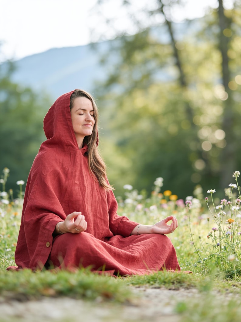 May include: A long-sleeved, hooded garment in a deep rust-red color, featuring a loose, flowing silhouette and a single visible wooden button on the side cuff. The fabric has a visible textured weave, and the hood is draped over the head.