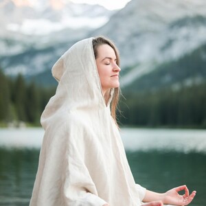 May include: A light beige hooded poncho made of a textured fabric. The garment features a loose, draped fit with a hood, three-quarter length sleeves, and a visible brown button detail near the hemline, providing a simple and functional design.