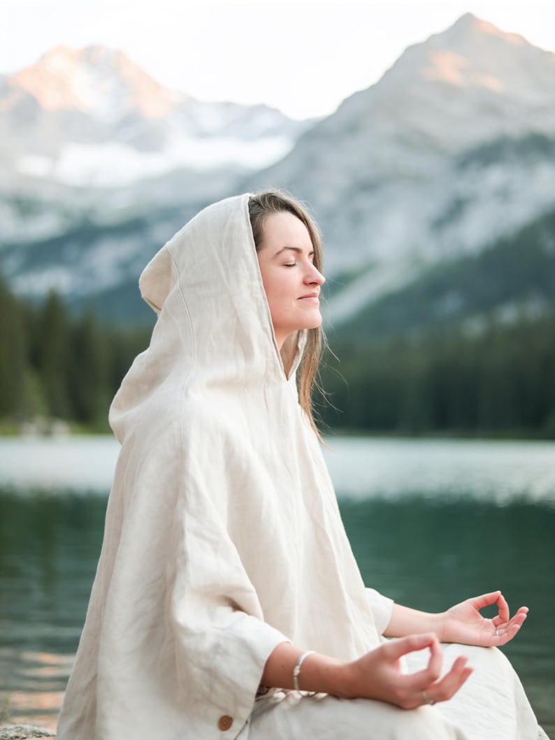 May include: A light beige hooded poncho made of a textured fabric. The garment features a loose, draped fit with a hood, three-quarter length sleeves, and a visible brown button detail near the hemline, providing a simple and functional design.